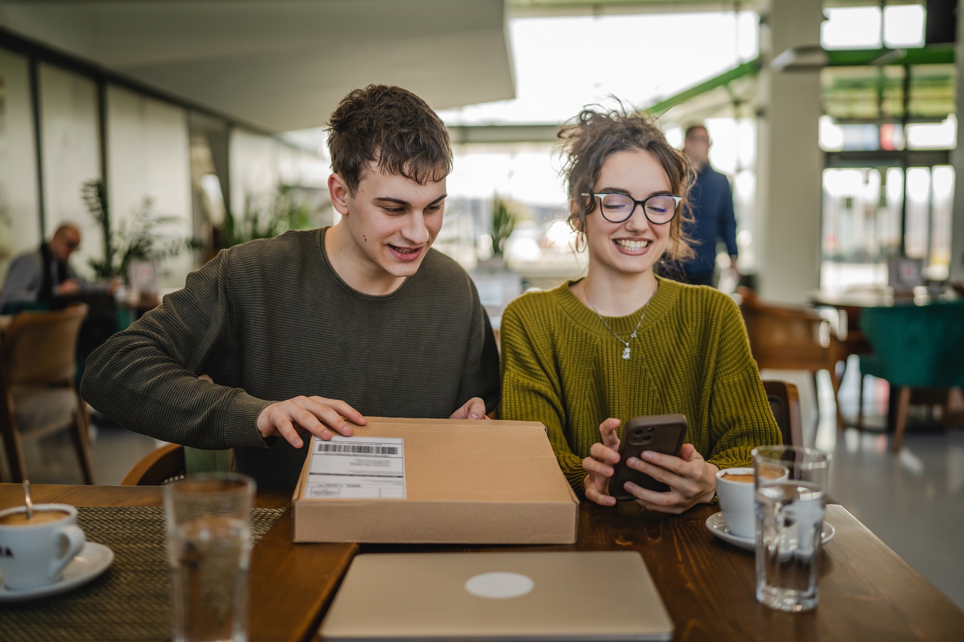 Gemini said Two students sitting in a university cafe smile while opening a cardboard delivery package