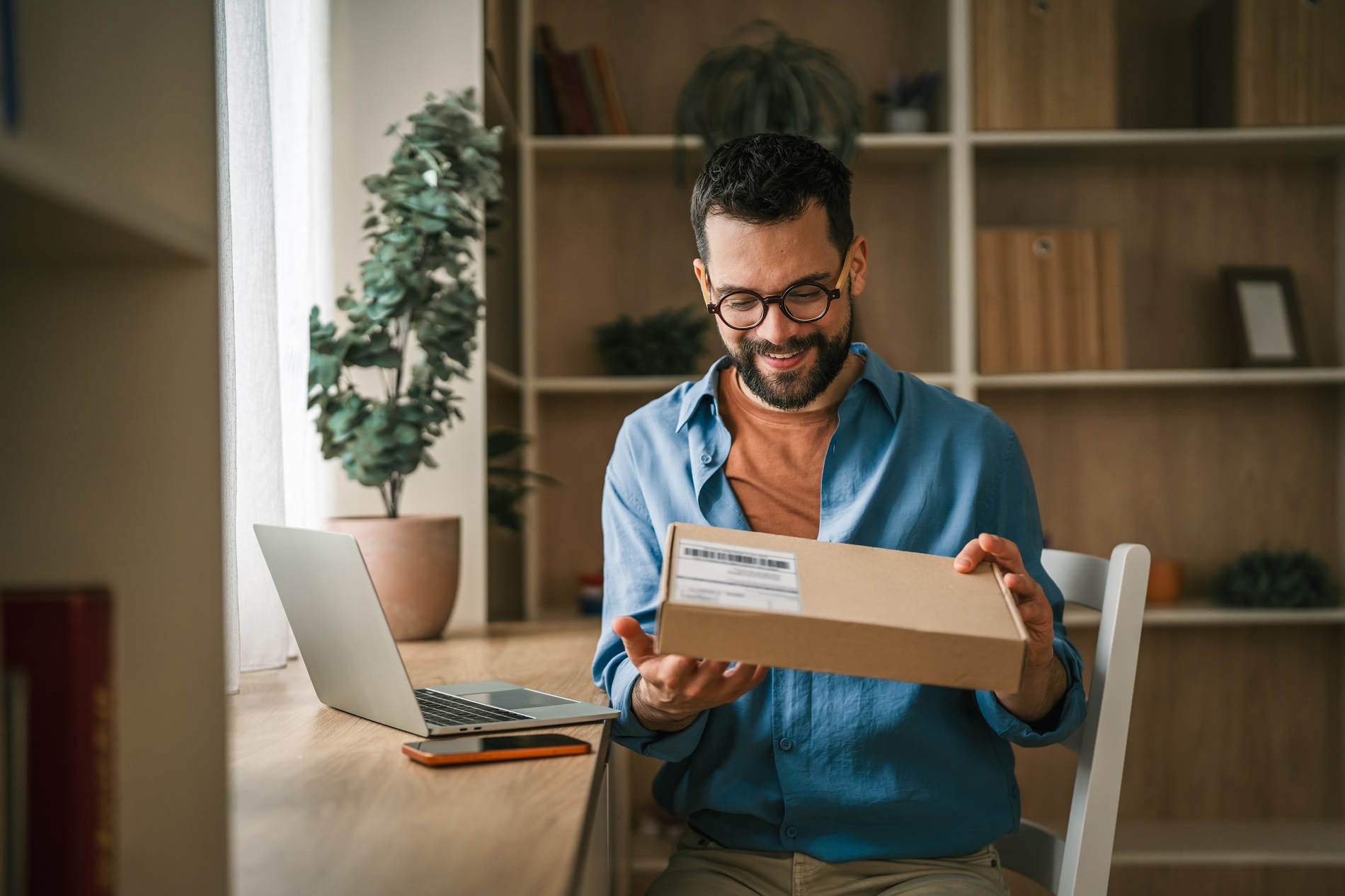 A smiling man sitting at a desk with a laptop and opening a delivered package
