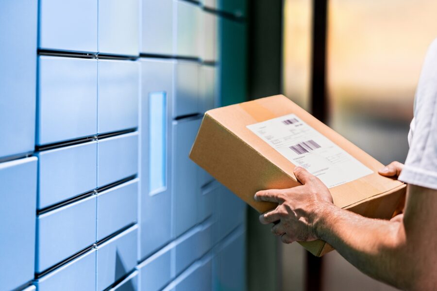 A person holding a cardboard delivery package in front of a bank of blue automated parcel lockers.